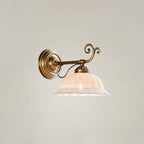 A Clérachic wall light with a straight brass arm and decorative scroll holds a small, lit white ceramic shade against a pale wall.