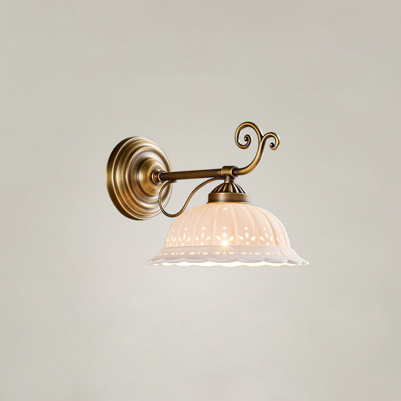 A Clérachic wall light with a straight brass arm and decorative scroll holds a small, lit white ceramic shade against a pale wall.