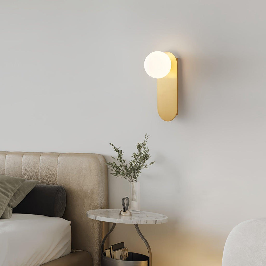 Lit brass globe sconce from Clérachic on a grey wall above a bedside table with a marble top and a sprig of olive.
