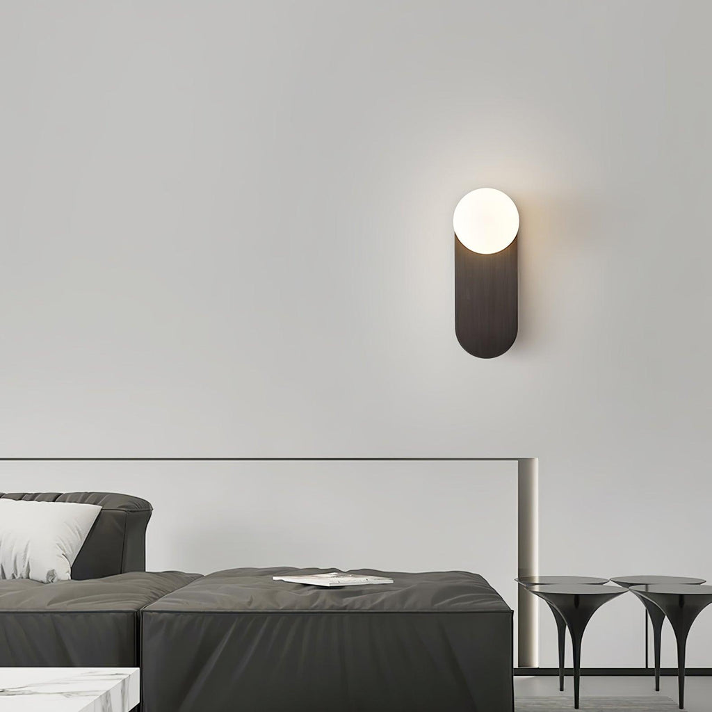Black Clérachic wall sconce with a glowing white globe on a pale wall above a low dark sofa and slim side tables.