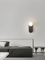 Small black-finish Clérachic wall light with a lit white globe above a dark ottoman and a pair of tripod tables.