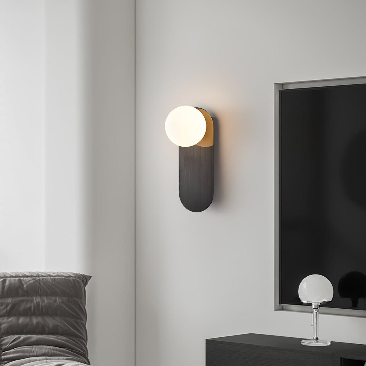 Black Clérachic globe sconce on a grey wall next to a dark screen, with a small glass-ball lamp on a console below.