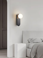 Black Clérachic globe sconce on a pale wall beside a tall dark door, with a white bed and bedside block below.