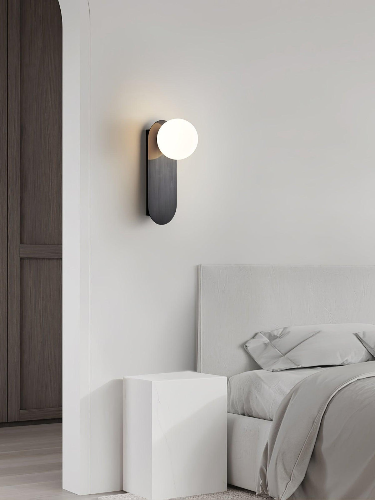 Black Clérachic globe sconce on a pale wall beside a tall dark door, with a white bed and bedside block below.