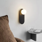 Lit black globe sconce from Clérachic above a floating bedside shelf with a glass and a book, next to a brown cushion.