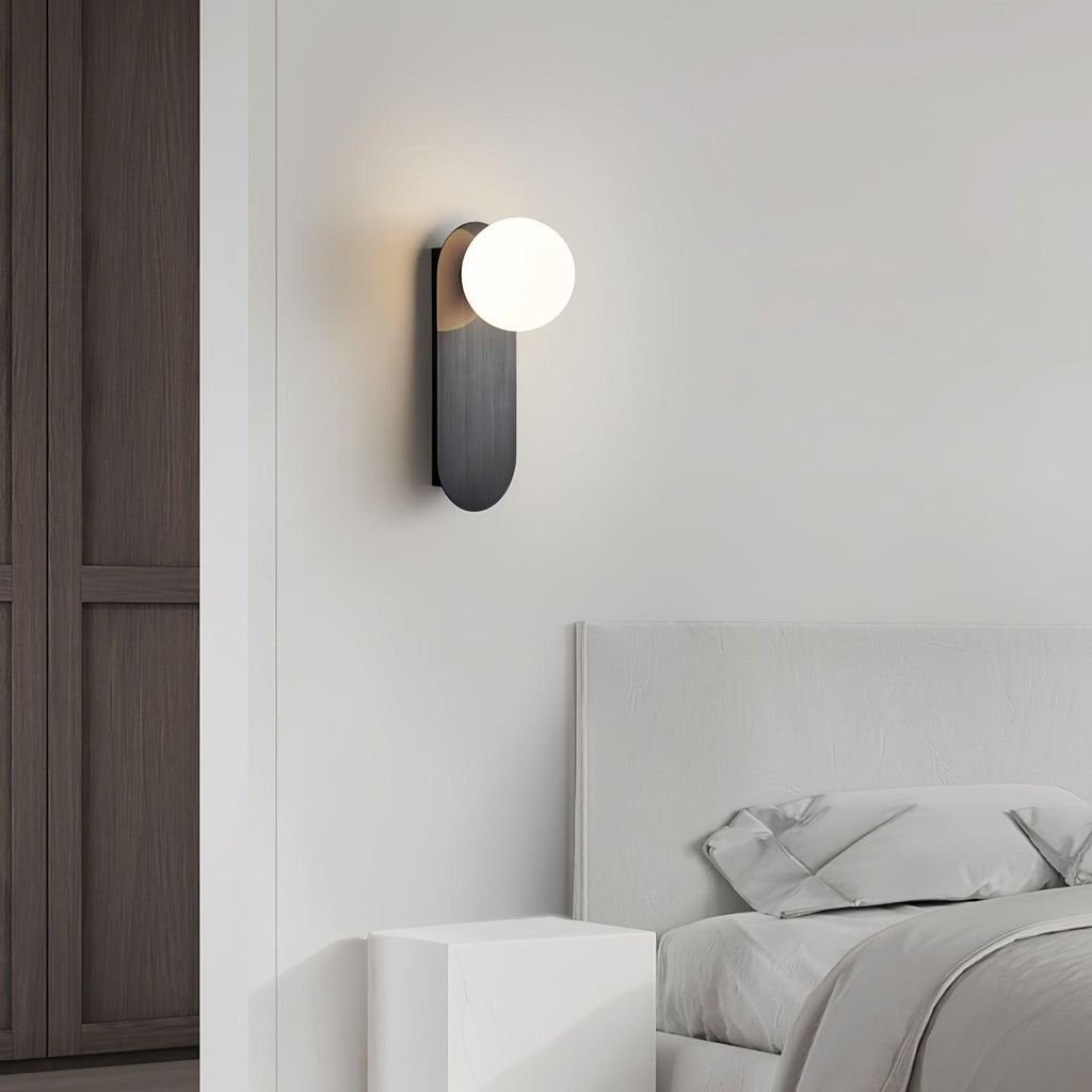 Black Clérachic globe sconce lit above a white upholstered bed, with a dark wood wardrobe door to one side.