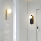 Brass and black Clérachic globe sconces on two white walls of a corridor, with a pale palm leaf and a door at right.