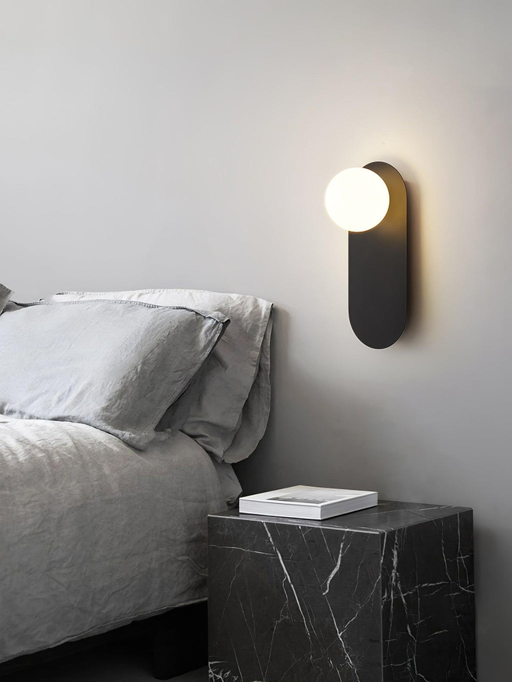 Black globe sconce from Clérachic above a bed with grey linen, next to a black marble bedside cube and a book.