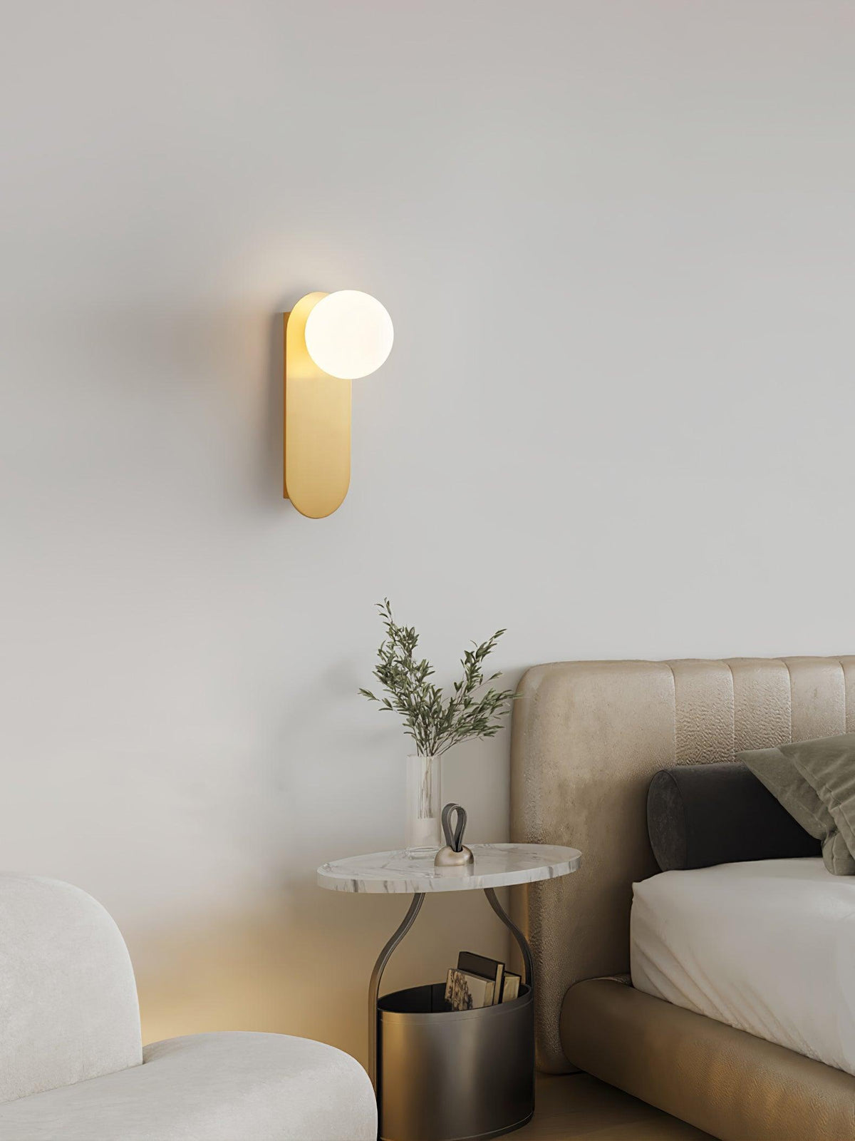 Lit brass globe wall light from Clérachic above a marble-topped side table with a plant, beside a cream bed and armchair.