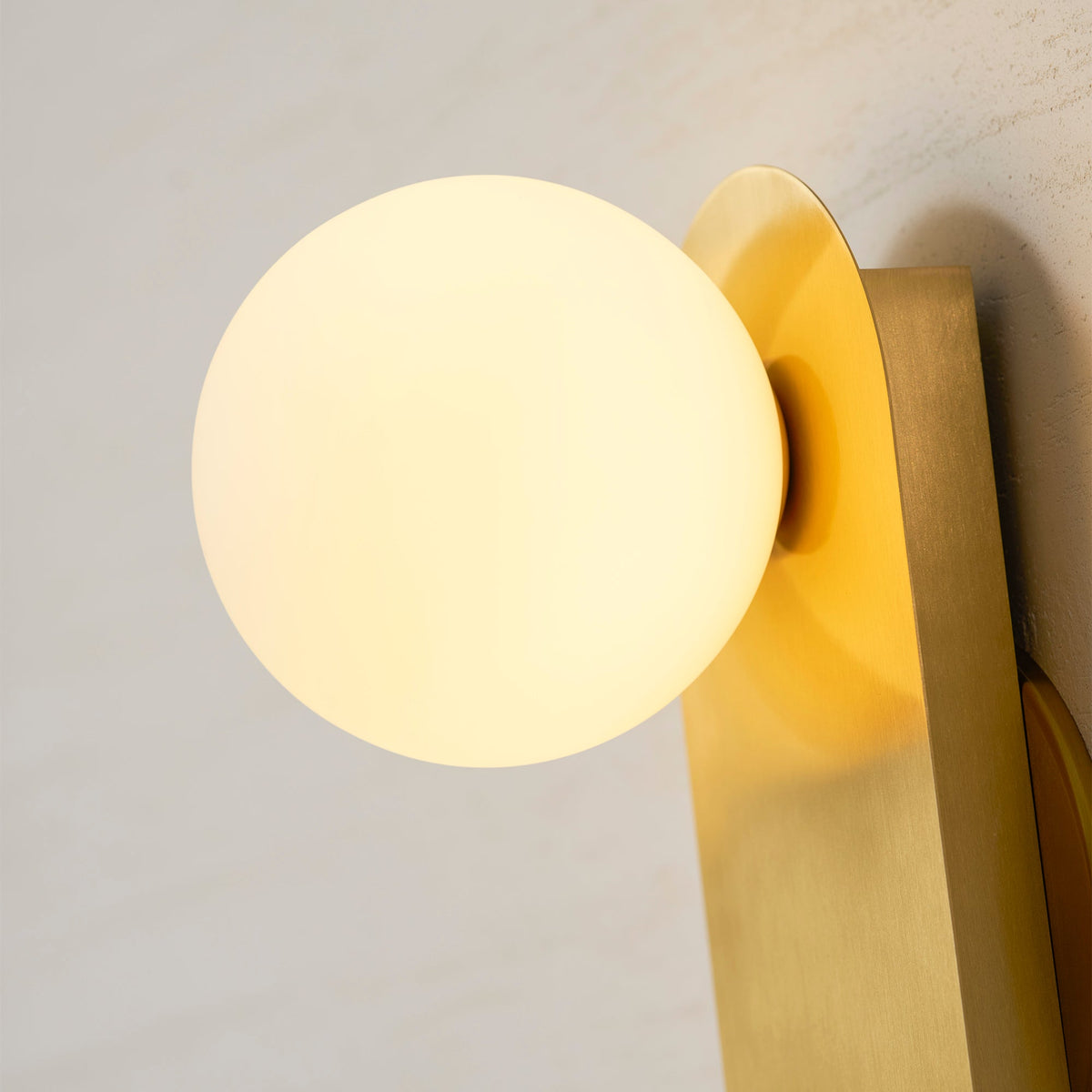 Close view of the glowing white globe and brushed brass plate on this Clérachic wall light, lit against a textured cream wall.