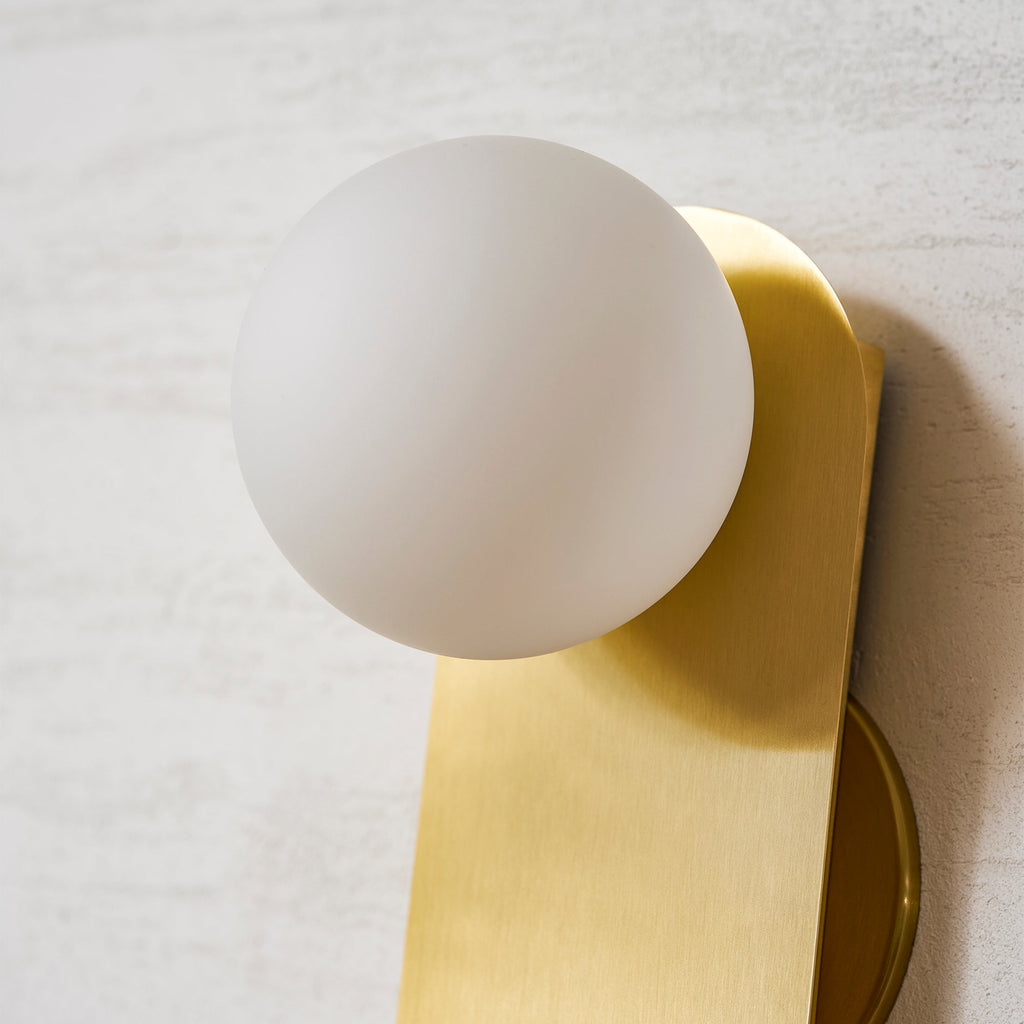 Close view of the unlit frosted white globe and brushed brass plate of this Clérachic wall light on a cream wall.