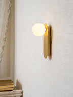 Lit brass globe sconce from Clérachic on a cream wall beside a pale curtain, with a stack of books on a shelf below.