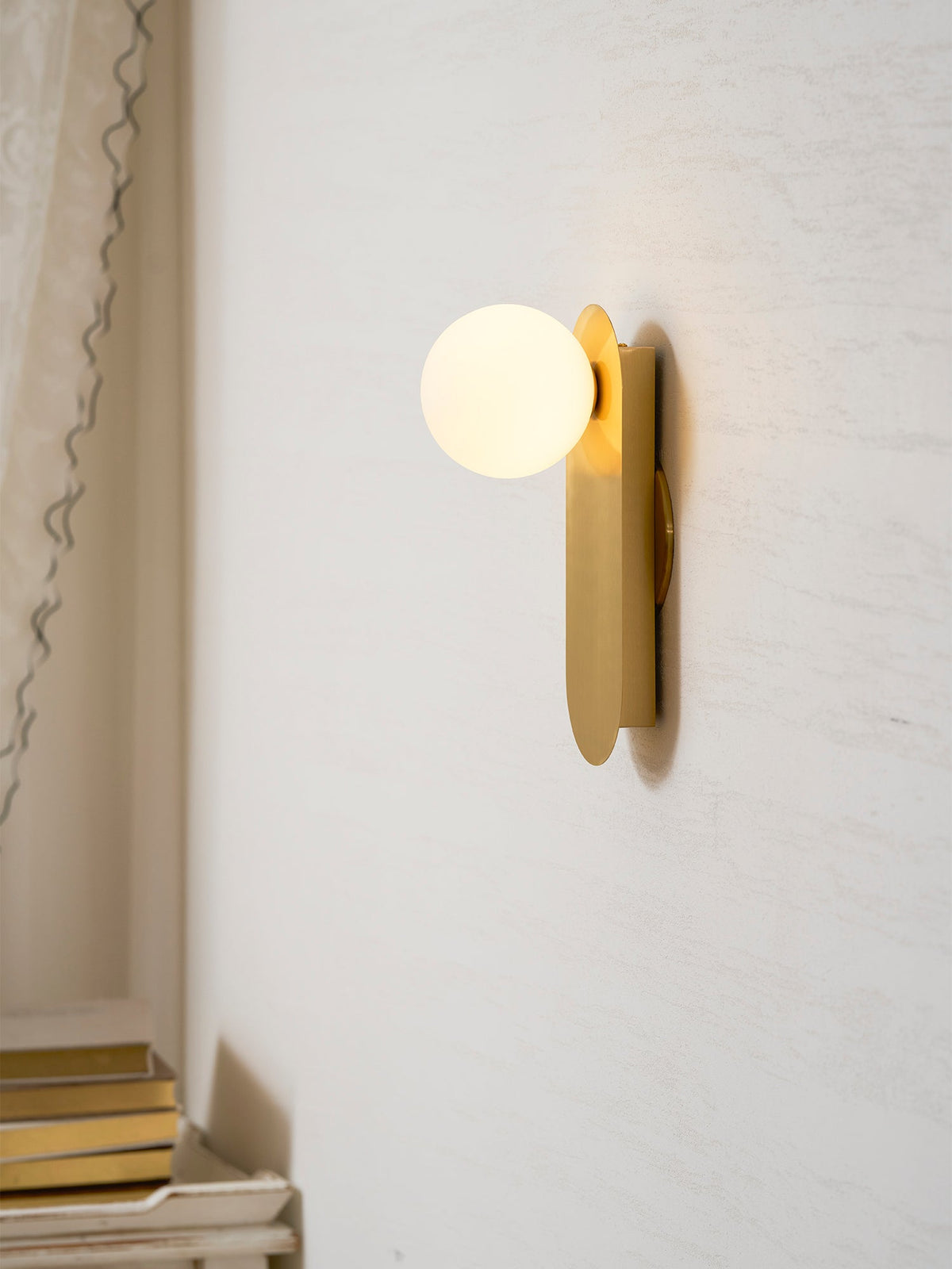 Lit brass globe sconce from Clérachic on a cream wall beside a pale curtain, with a stack of books on a shelf below.