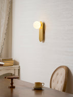 Small lit brass Clérachic wall light on a cream wall above a dark wood table, a cup and a button-back chair.