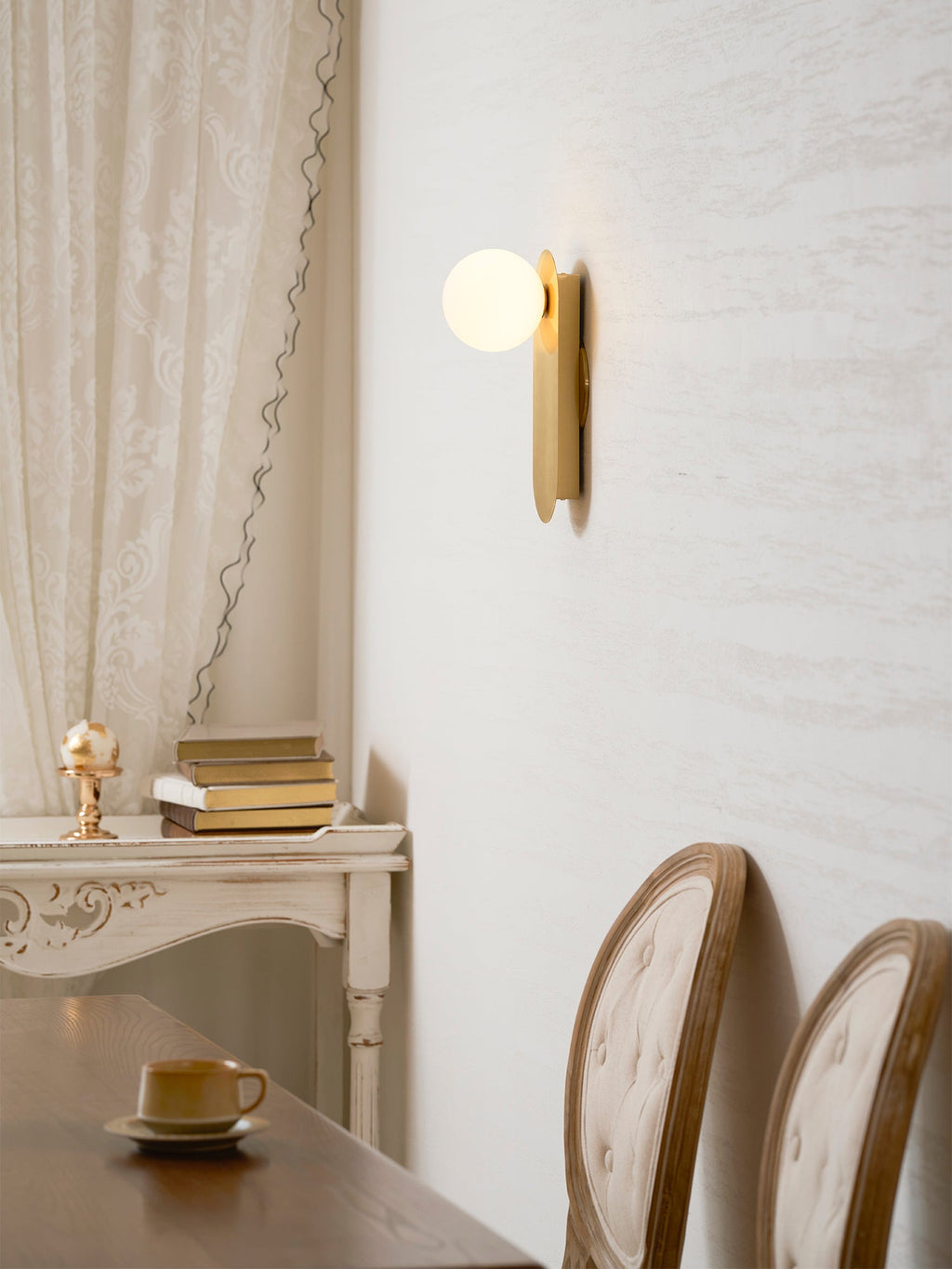 Brass globe sconce from Clérachic above a table set with a cup, with two button-back chairs and a lace curtain beside it.