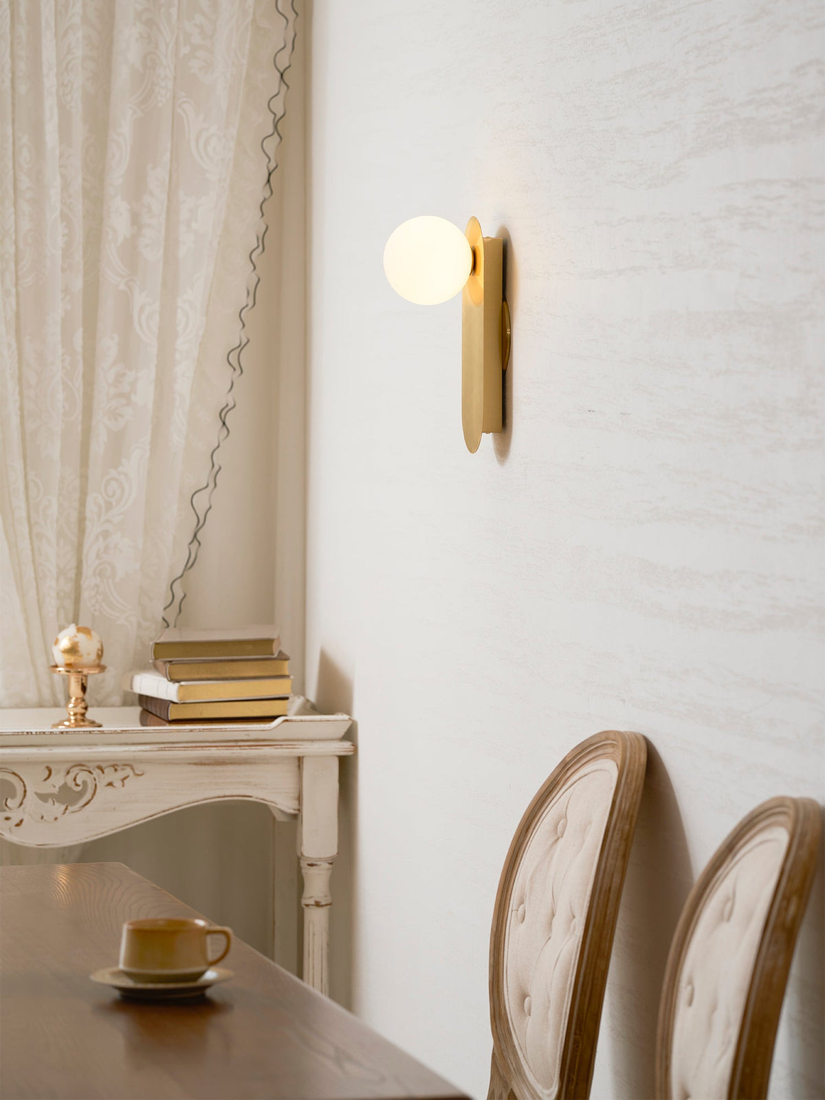 Brass globe sconce from Clérachic above a table set with a cup, with two button-back chairs and a lace curtain beside it.