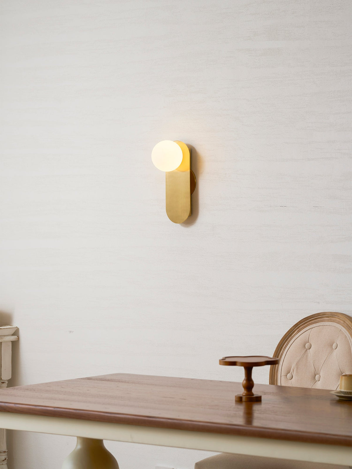Lit brass Clérachic globe wall light on a textured cream wall above a wooden table with a small cake stand.