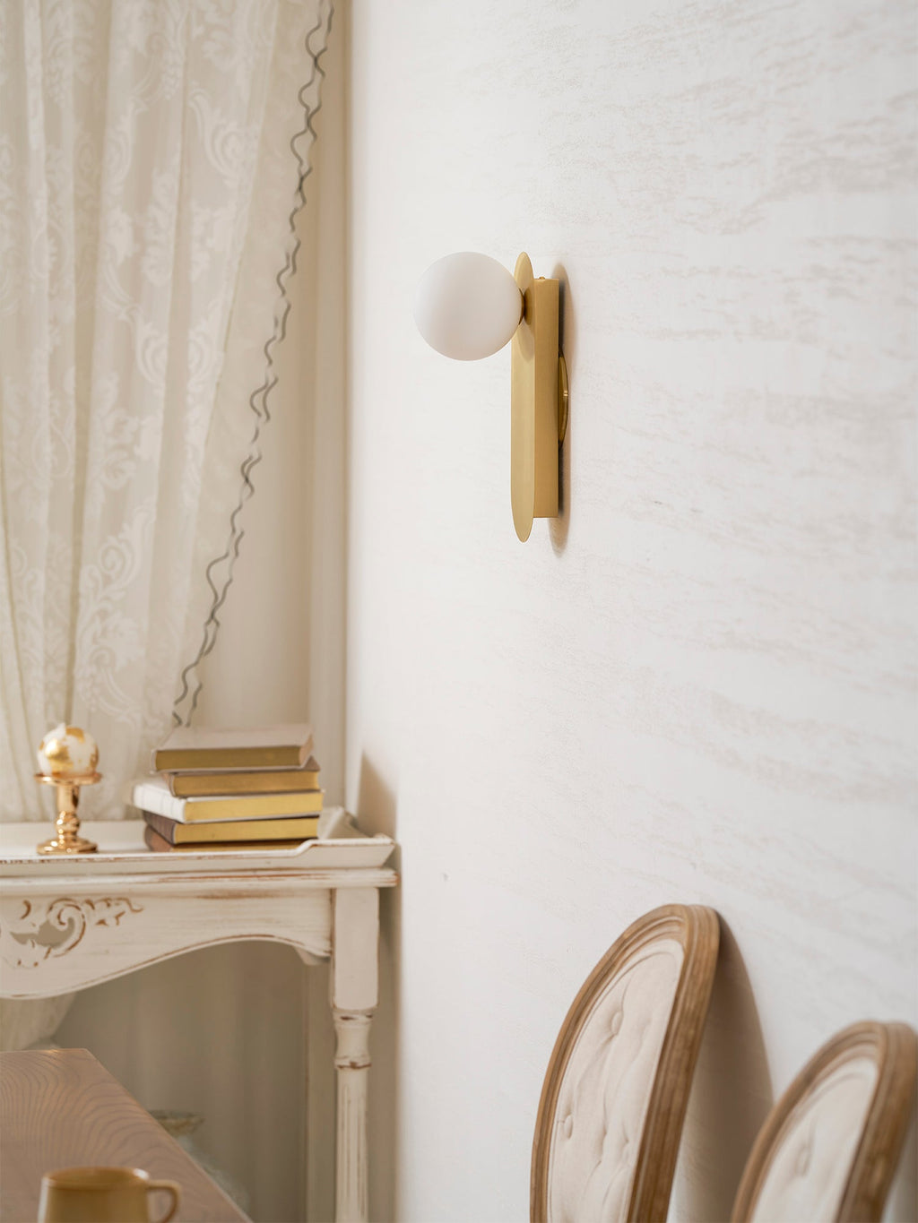 Unlit brass globe wall light from Clérachic beside a lace curtain, above a white console with books and a small ornament.