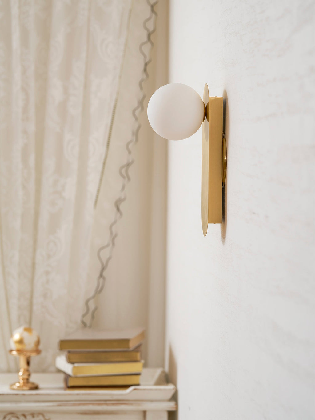 Close view of the white globe and brass plate of this Clérachic wall light, a lace curtain and a stack of books behind.