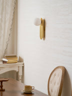 Brass globe wall light from Clérachic seen from a low angle on a cream wall, with a white console and a chair below.