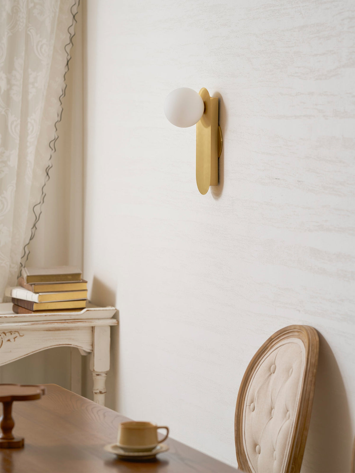 Brass globe wall light from Clérachic seen from a low angle on a cream wall, with a white console and a chair below.