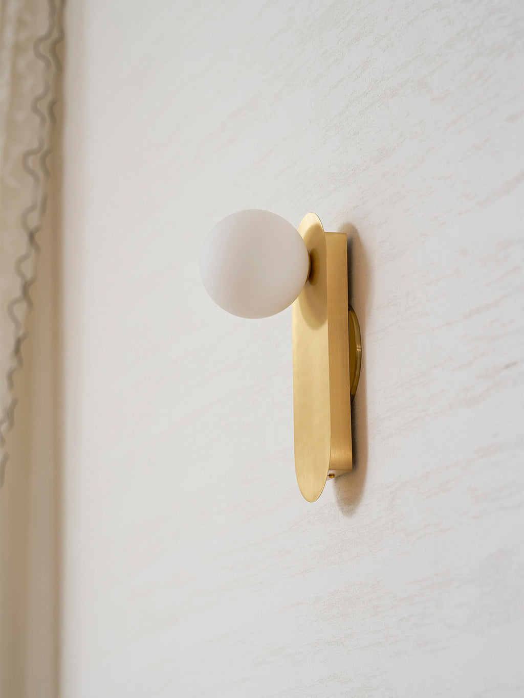 Side view of the brass plate and white globe, this Clérachic wall light close to a cream wall by a curtain edge.