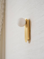 Side view of the brass plate and white globe, this Clérachic wall light close to a cream wall by a curtain edge.