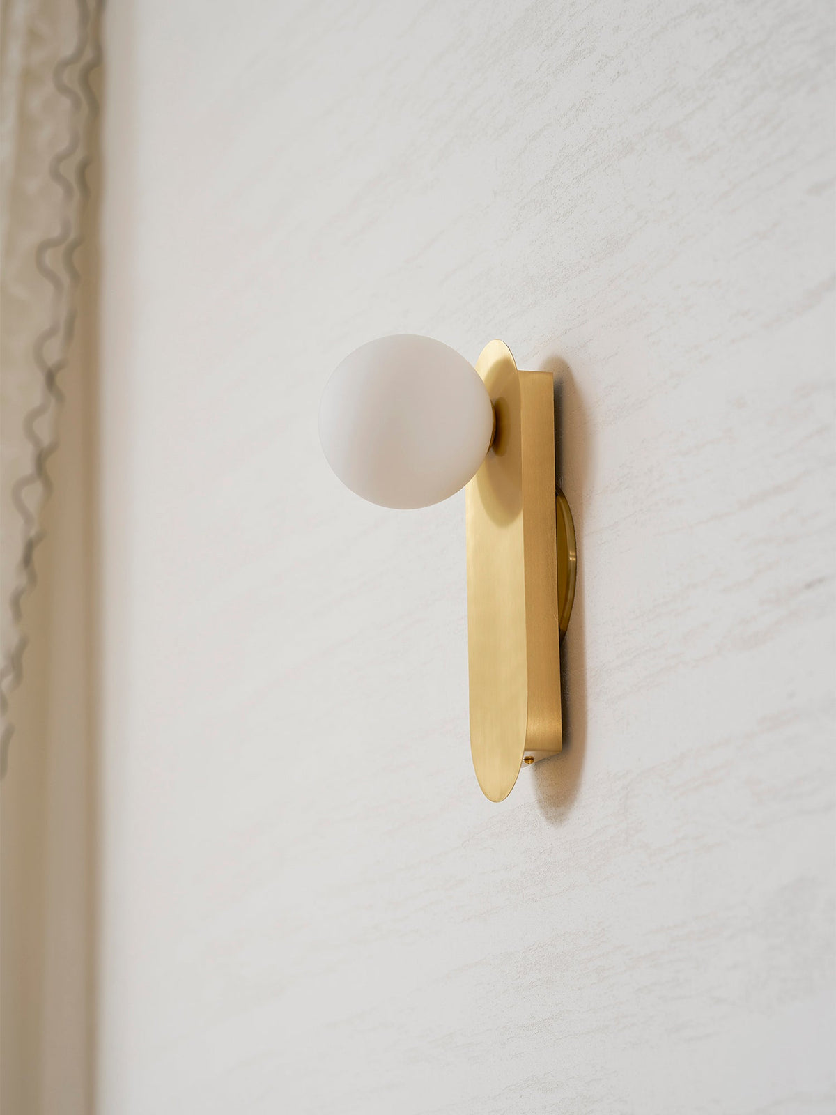 Side view of the brass plate and white globe, this Clérachic wall light close to a cream wall by a curtain edge.