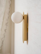 Close side view showing how the white globe projects from the brass plate of this Clérachic wall light, a lace curtain behind.