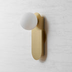 Unlit white globe and brushed brass plate of this Clérachic wall light, at a slight angle against a grey textured wall.