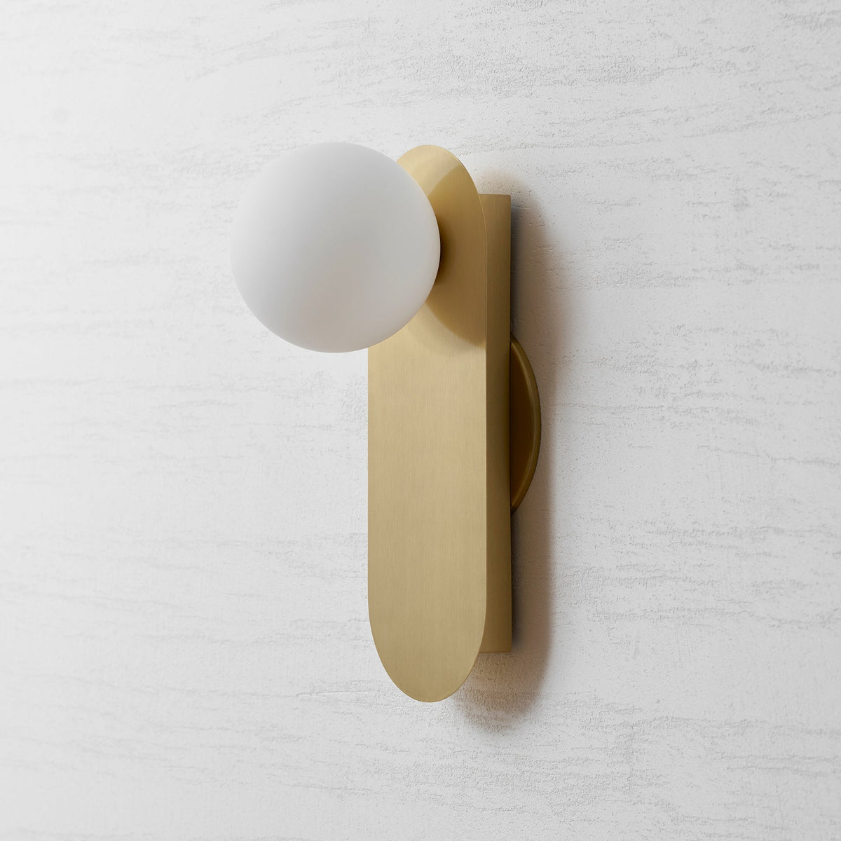 Unlit white globe and brushed brass plate of this Clérachic wall light, at a slight angle against a grey textured wall.