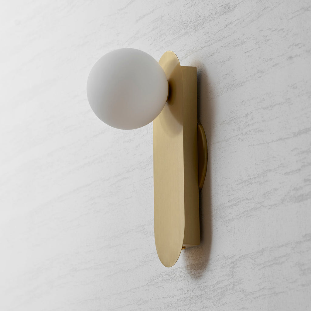 Side-on view of the brushed brass plate and frosted white globe of this Clérachic wall sconce on a grey textured wall.