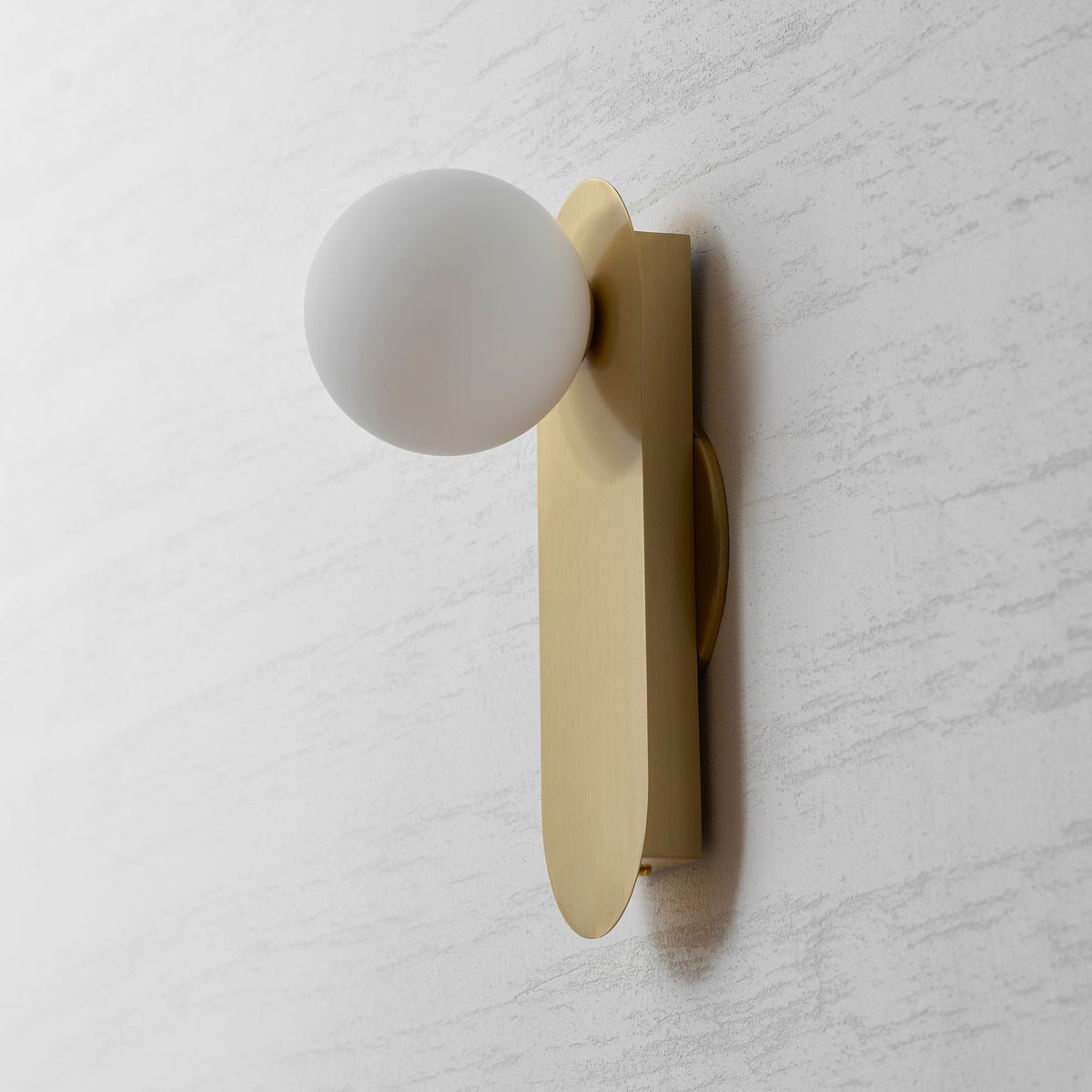 Side-on view of the brushed brass plate and frosted white globe of this Clérachic wall sconce on a grey textured wall.