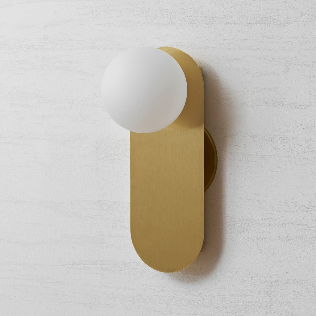 Three-quarter front view of this Clérachic brass wall light, the white globe overlapping the top of the rounded plate.