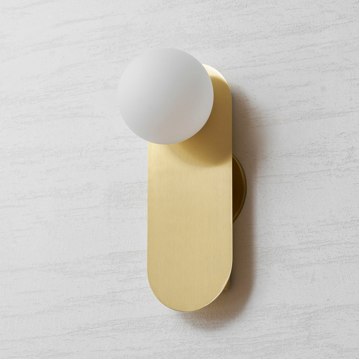 Brass wall light from Clérachic with a frosted white globe, photographed slightly from the right on a textured wall.