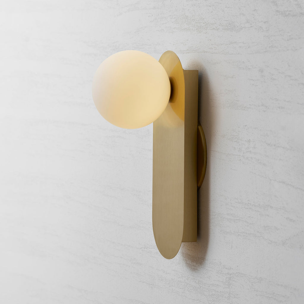 Angled view of the brass plate, its thin edge and the white globe of this Clérachic wall light on a grey wall.