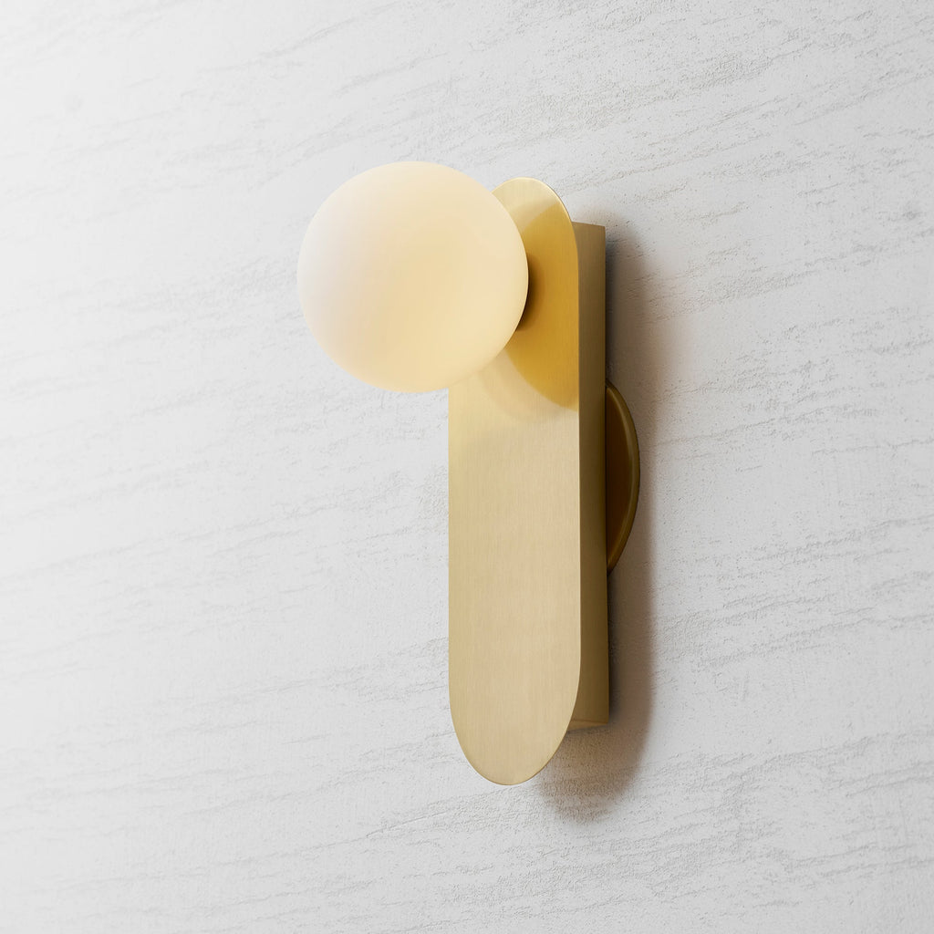 Brass globe wall light from Clérachic seen from the left, the plate edge and the globe's soft glow visible on the wall.