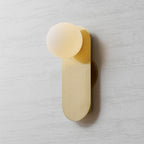 Clérachic brass wall light with a warm-glowing white globe, shown slightly from the left against a pale textured wall.