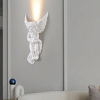 The cherub version of this Clérachic wall light, lit, sending a column of warm light up a grey panelled wall above a cream sofa.