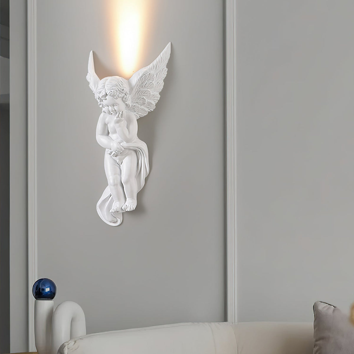 The cherub version of this Clérachic wall light, lit, sending a column of warm light up a grey panelled wall above a cream sofa.