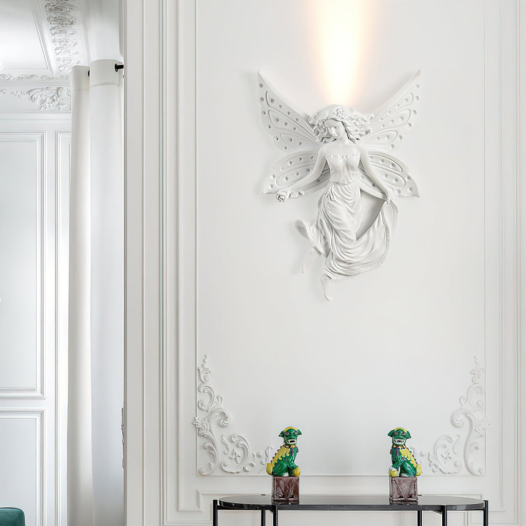 Lit fairy-shaped Clérachic wall light on a white moulded wall, with two small green figurines on a dark console below.