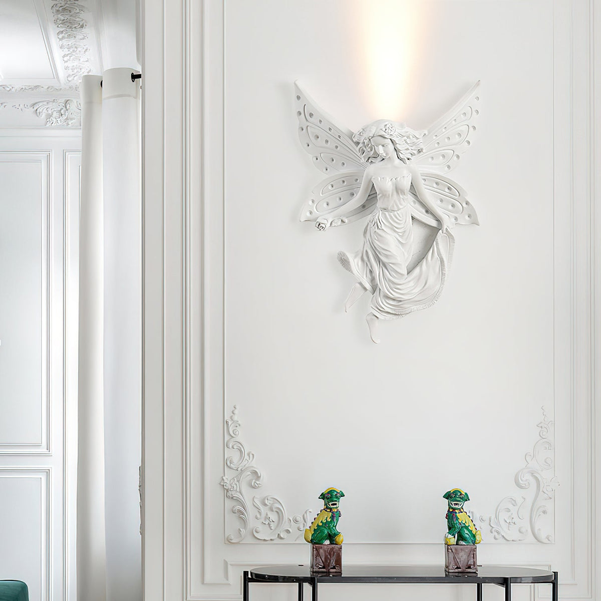 Lit fairy-shaped Clérachic wall light on a white moulded wall, with two small green figurines on a dark console below.