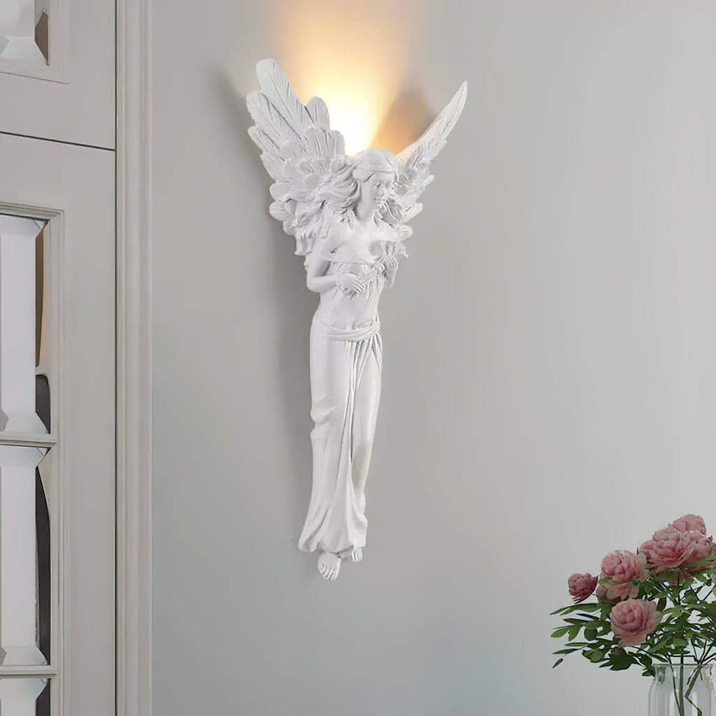 Tall, lit angel-shaped Clérachic wall light on a pale grey wall, with a bunch of pink roses at the lower right.