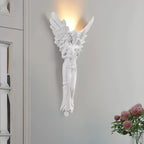 Tall, lit angel-shaped Clérachic wall light on a pale grey wall, with a bunch of pink roses at the lower right.