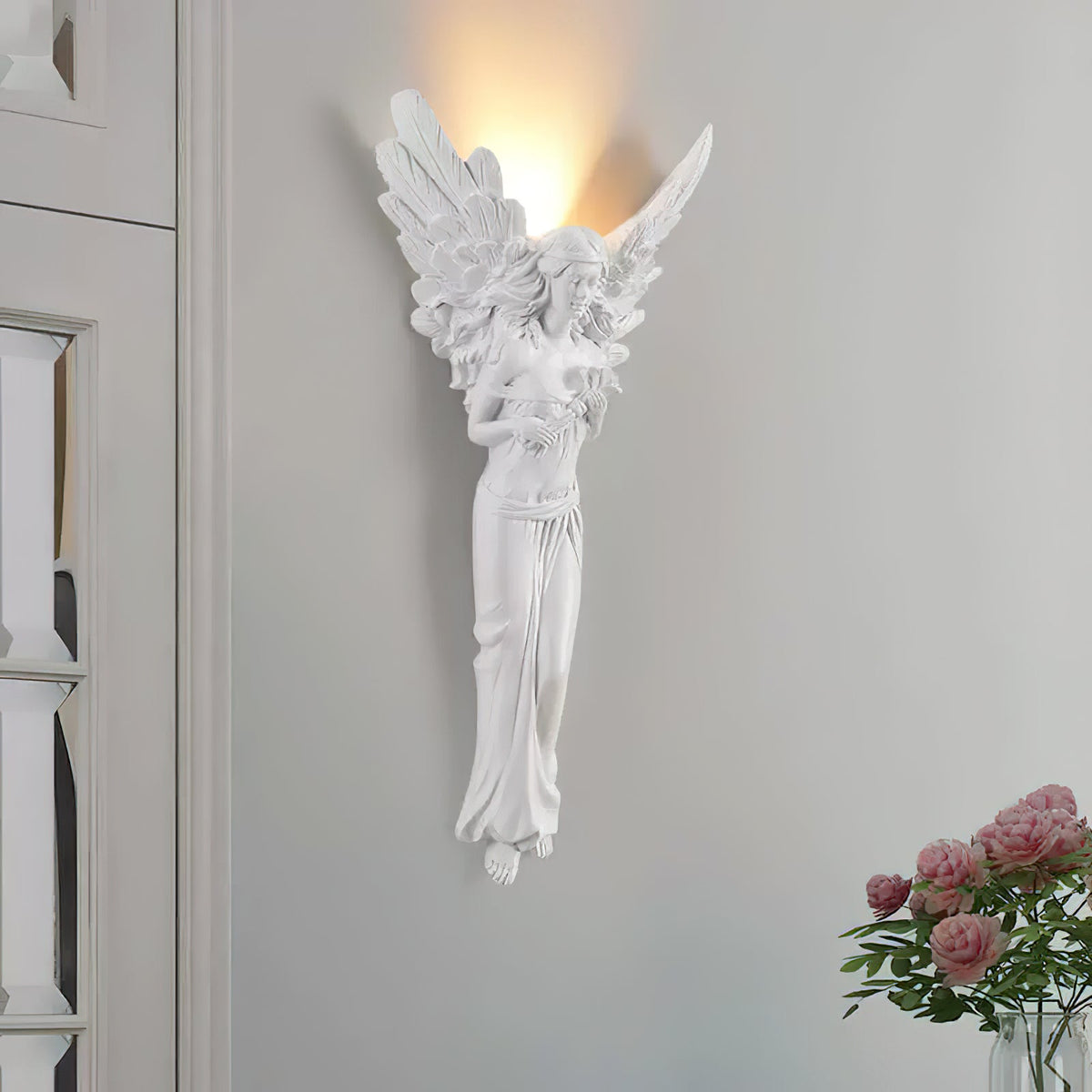 Tall, lit angel-shaped Clérachic wall light on a pale grey wall, with a bunch of pink roses at the lower right.