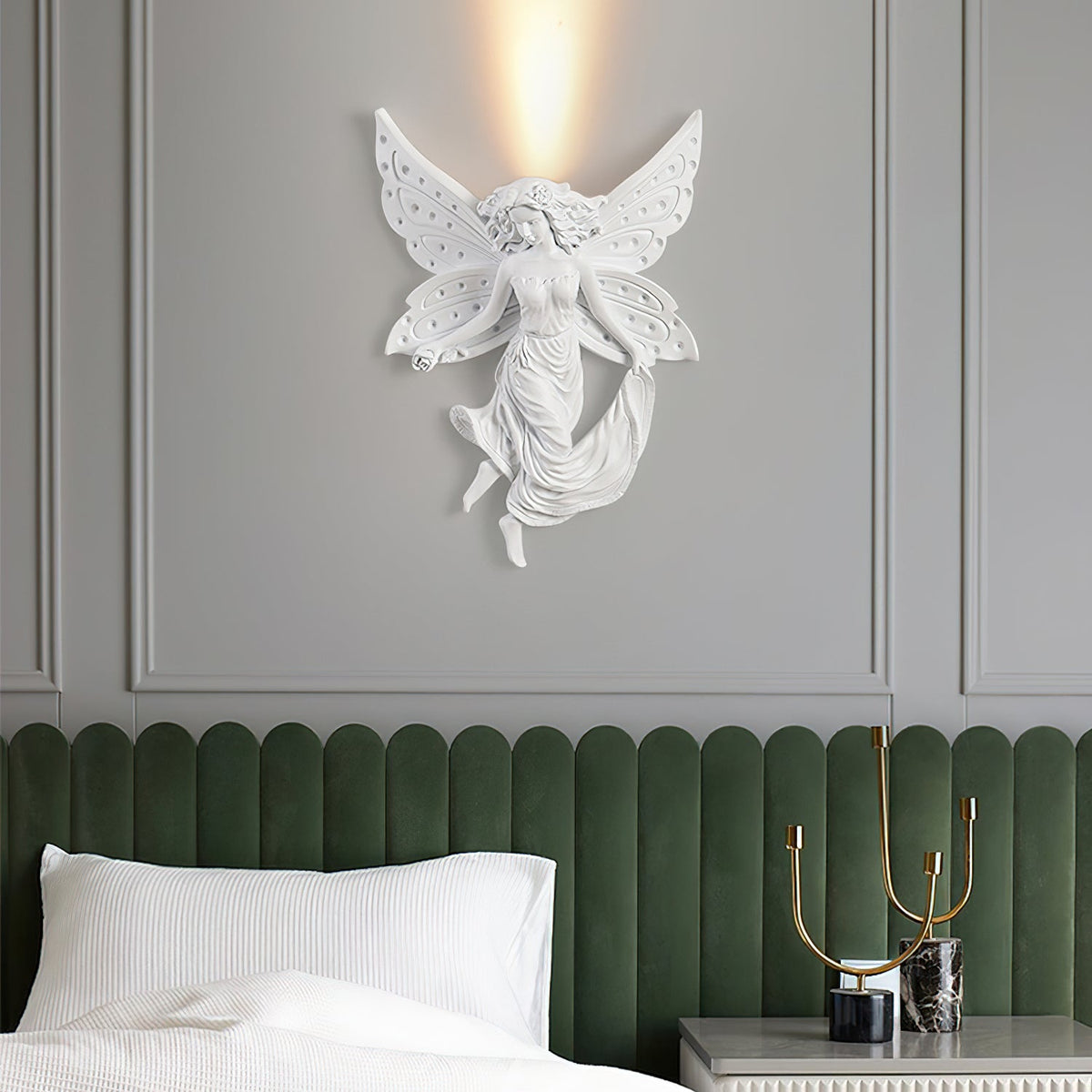 Fairy-shaped Clérachic wall light, lit, on a grey wall above a green headboard, a white pillow and a gold candle holder.