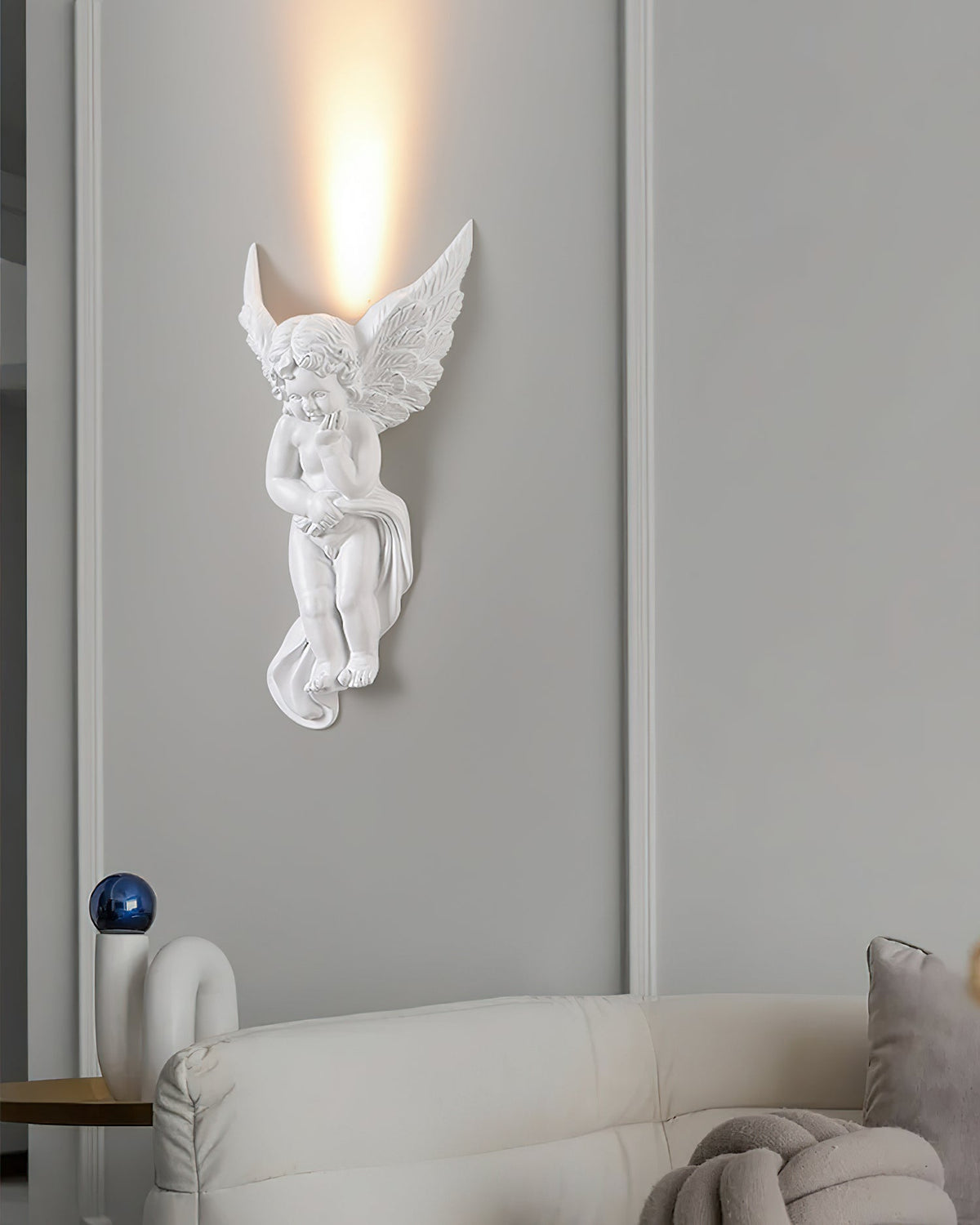 Cherub-shaped Clérachic wall light lit on a tall grey panelled wall, a cream sofa and a grey knitted cushion below.