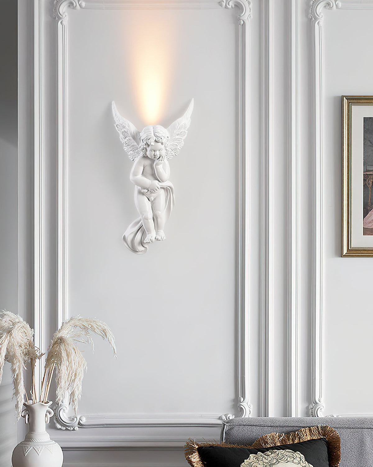 Cherub Clérachic wall light lit on a white moulded wall, with a vase of dried pampas grass at the lower left.
