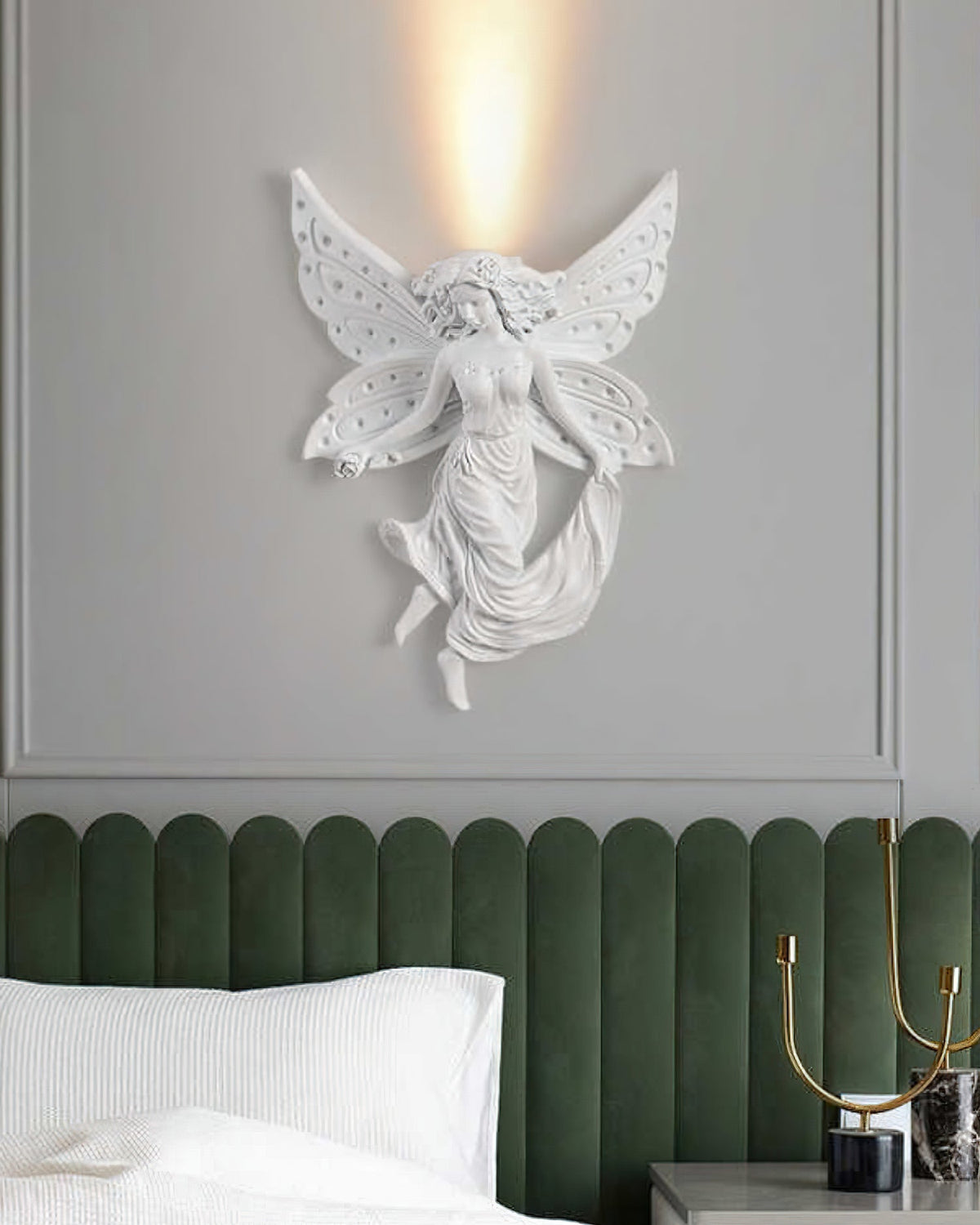 A Clérachic fairy wall light, lit, on a grey wall with the warm uplight rising from behind its wings, above a green headboard.