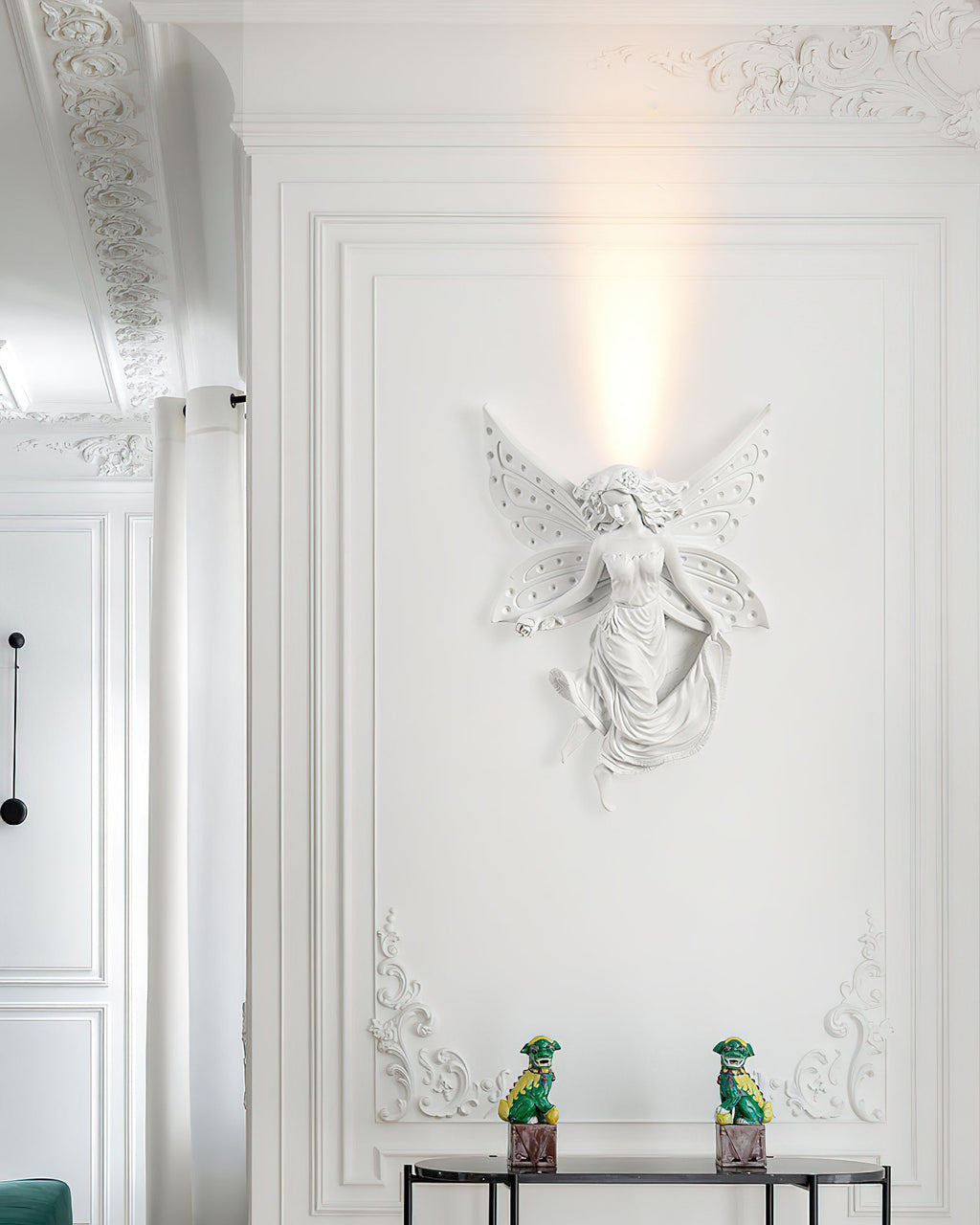 Fairy-shaped Clérachic wall light lit on a tall white wall with ornate plasterwork and two small green figurines on a console.
